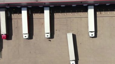 Semi truck with cargo trailer is travelling along a parking lot of a warehouse in the logistics park. A lot of semi-trailers trucks stands at warehouses ramps for load/unload goods. Aerial view
