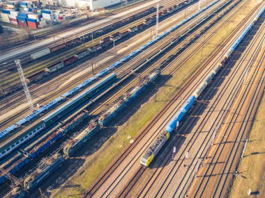 Kargo trenleri. Renkli yük trenlerinin hava manzarası. Tren istasyonunda. Demiryolunda malları olan vagonlar. Ağır endüstri. Trenlerle, şehir binalarıyla ve gün batımında bulutlu gökyüzüyle sanayi manzarası. Üst görünüm