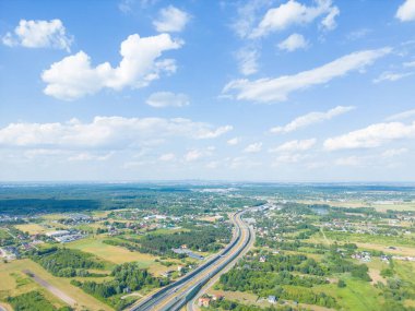 Yazın günbatımında yeşil çayırlardaki yol manzarası. Kırsal yol ve ormanın insansız hava aracı manzarası. Yol, güneş ışınları, ağaçlar, tepeler, yeşil çimenler, bulutlar olan güzel bir manzara. Polonya