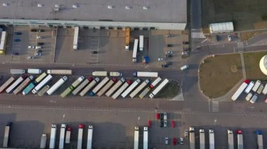 Semi truck with cargo trailer is travelling along a parking lot of a warehouse in the logistics park. A lot of semi-trailers trucks stands at warehouses ramps for load/unload goods. Aerial view