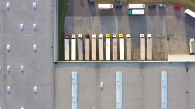 Semi truck with cargo trailer is travelling along a parking lot of a warehouse in the logistics park. A lot of semi-trailers trucks stands at warehouses ramps for load/unload goods. Aerial view