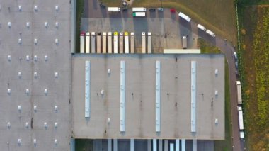 Semi truck with cargo trailer is travelling along a parking lot of a warehouse in the logistics park. A lot of semi-trailers trucks stands at warehouses ramps for load/unload goods. Aerial view