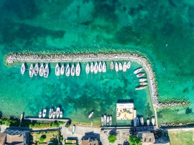 Fransa 'nın Yvoire limanındaki bir turkuaz su limanında hilal şeklinde demirli beyaz yelkenlilerin havadan aşağı görüntüsü. 