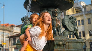 Güzel kızıl saçlı anne ve küçük kız oyun oynuyorlar, eski Avrupa kasabasındaki çeşmenin yanında eğleniyorlar..