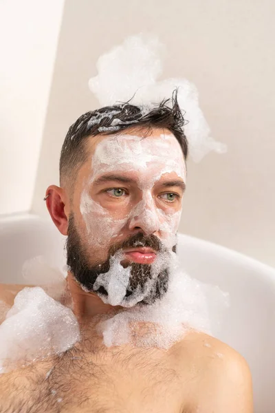 Funny Bearded man with white mask on face ans soap form on the head in bath at home