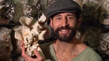 Kameraya gülümseyen ve istiridye mantarı çiftliğine karşı mantar tutan mutlu genç adam. Organik tarım tarım çiftliği..