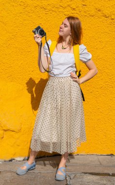 young tourist red haired woman holding a camera on colorful background wall in summer. full length body shot