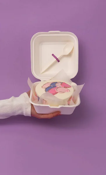 Hand holding Bento mini cake as a gift for the holiday on minimalistic purple paper background. Korean style cakes in a box for one person