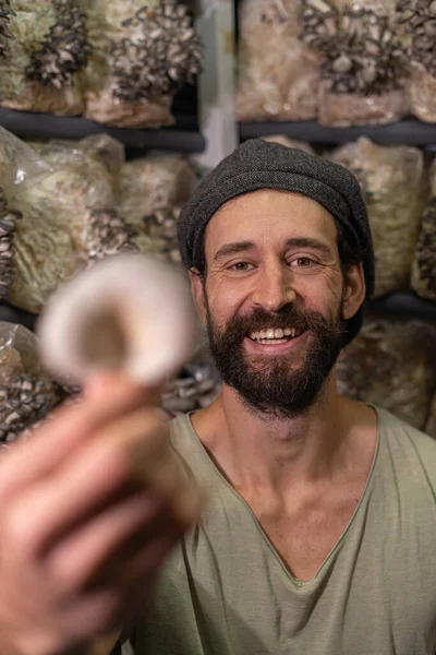 happy young man smiling to camera and holding mushroom against the oyster mushrooms farm, selective focus. Organic plantation cultivation farm business.