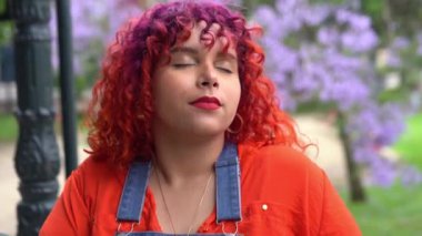 Renkli saçlı genç bir kadın gözlerini kapar ve şehir parkındaki Jacaranda çiçeklerinin kokusunu alır. Yakın plan.