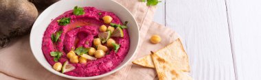 Homemade Beetroot hummus decorated with chickpeas on pink towel, pita bread and with fresh beetroots on the white wooden table