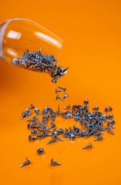 Bir sürü metal vidayı bir bardakta, kova kavanozunda düşürdüm. Metal için kendi kendine dokunan vida, demir, krom kaplama kendi kendine dokunabilen vida, turuncu sarı bir arka planda, metin için boşluk