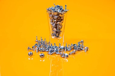 Bir sürü metal vida, bir bardakta, kova kavanozunda çelikten yapılmıştır. Metal için kendi kendine dokunan vida, demir, krom kaplama kendi kendine dokunabilen vida, turuncu sarı bir arka planda, metin için boşluk