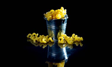 Cam kavanozda çiğ makarna, şarap kadehi. Kovayla. Siyah arka planda çiğ makarna. Ön manzara ham makarna, elden düşme, yazı için yer, sarı uzun spagetti pipetleri, yiyecek malzemeleri.