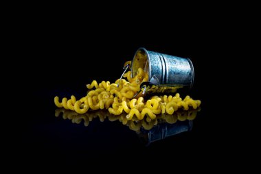 Cam kavanozda çiğ makarna, şarap kadehi. Kovayla. Siyah arka planda çiğ makarna. Ön manzara ham makarna, elden düşme, yazı için yer, sarı uzun spagetti pipetleri, yiyecek malzemeleri.