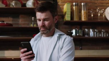 Bir adam arkasında şık ve modern bir mutfak zemininde dururken akıllı telefonuyla meşgul olur.