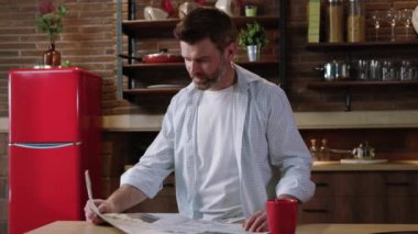 Yakışıklı, orta yaşlı bir adamın kahvaltıda mutfakta dinlenirken çekilmiş görüntüleri. Şaşırmış bir adamın portresi gazetede heyecan verici komik haberler okuyor ve masanın başında gülümsüyor..