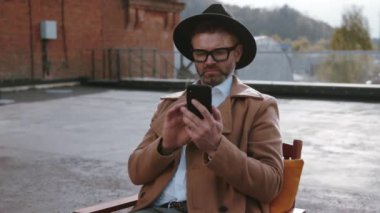 Şapkalı ve ceketli şık bir erkek model terasta sandalyede oturmuş dışarıda sohbet etmek için akıllı telefon kullanıyor..