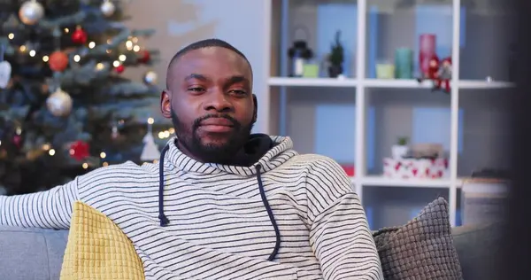 Kahverengi gözlü genç Afrikalı adam Noel ağacının arka planında topları ve süslemeleri ile dikkatle izliyor. Yakın plan..