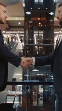 Meşgul bir alışveriş merkezinde, iki uygun adam profesyonel etkileşim içinde, çeşitli ortamlarda tokalaşarak iş görüşmelerini sergiliyor, güvenlerini yansıtıyor.