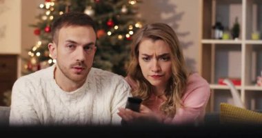 Kız ve erkek televizyon izleyip tartışıyorlar. Genç ve güzel bir kadın dekore edilmiş Noel ağacının arka planında televizyon kanallarını değiştiriyor. Kutlama atmosferi. Kapalı.