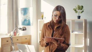 Oturma odasında masanın yanında duran gözlüklü güzel Ukraynalı kadın. Tableti elinde tutan, makale okuyan bir kız. Genç kadın kameraya bakıyor ve gülümsüyor..