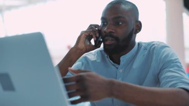 Ciddi bir Afro-Amerikan genci kafeterya portresinde dizüstü bilgisayar ve konuşma telefonu kullanıyor. Sadece yüz teknolojisini kapat. Siyah, portatif, çekici bir adam sıradan bir hayat tarzı telefon yakışıklı masasında oturuyor.