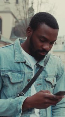 Sıradan bir kot ceket giymiş genç bir adam dışarıda şehir ortamında akıllı telefonunu kullanıyor.