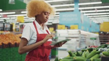 Güzel Afro-Amerikalı kadın satıcı, süpermarkette sebze reyonunda çalışmak için tablet kullanıyor. Markette çalışan, bakkal, sağlıklı personel, perakende satış mağazası üretiyor.