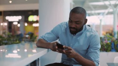 Portre gülümseyen Afrikalı Amerikalı genç adam telefon mesajı kullanıyor alışveriş merkezindeki masada oturuyor mutlu, siyah telefon kapatıyor ağır çekimde sevimli cep telefonu tutuyor.