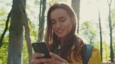Ormanın ortasında parlak bir güneş ışığında sırt çantalı, mutlu bir şekilde gülümseyen ve akıllı telefonunu kullanan esmer bir kadının dönüşümlü görüntüsü. Sosyal ağlar, aygıtlar, aygıtlar.