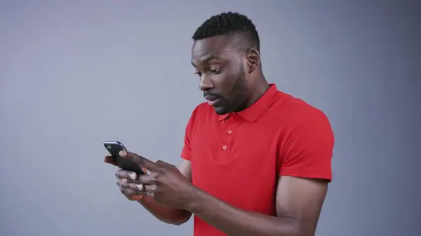 Yakışıklı, sakallı, Afro-Amerikan bir adam gri arka planda mesajlaşmak için akıllı telefonunu aktif olarak kullanıyor, şaşırıyor ve tatlı tatlı gülümsüyor. İletişim, çevrimiçi olmak, sohbet etmek. Erkek portresi