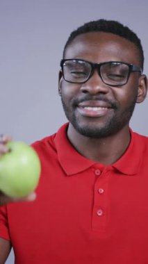 Kırmızı polo tişörtlü yakışıklı Afro-Amerikalı adam gri arka planda gülümseyen taze yeşil elmayı kameranın önünde tutuyor. Vitaminler, formda olmak, diyet konsepti. Erkek portresi, yakın görünüm