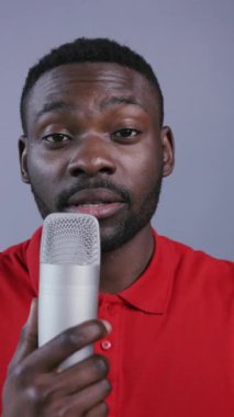 Kırmızı polo tişörtlü neşeli Afro-Amerikalı adam mikrofon kullanıyor ve gri arka planda konuşuyor. Stand-up komedyeni, şaka yapıyor, eğleniyor, kayıt yapıyor. Erkek portresi, yakın görünüm