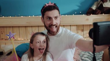 Yakın plan portre. Komik baba ve kızı küçük prenses. Dilleri buruşuk, evde, yatak odasında ortak selfie çekiyorlar..