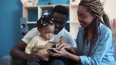 Genç Afro-Amerikan ebeveynler sevimli fiyonklu ve oyuncak ayılı tatlı kızlarına akıllı telefonu nasıl kullanacaklarını öğretirler. Aile zamanı, modern çocukluk. Görüntüyü kapat, kamera sabitleyici.