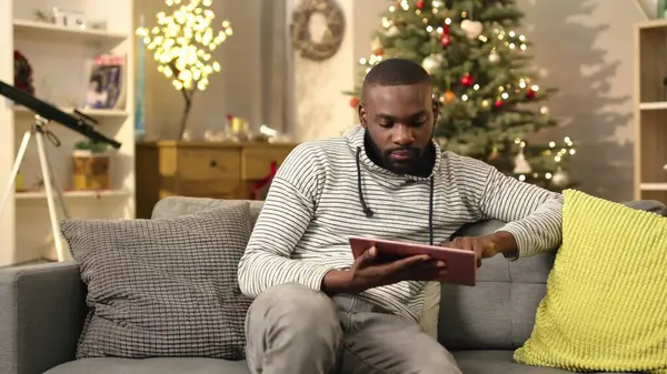 Kanepede oturan ve dijital tableti elinde tutan yakışıklı bir Afrikalı. Genç adam internette bir şeyler arıyor. İçeride. Noel atmosferi.
