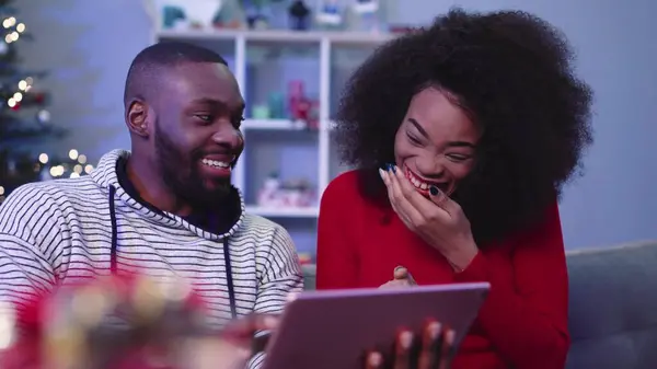 İki genç, güzel Noel ağacının arka planında tablet üzerinde oyun oynuyor. Çekici Afrikalı çift zafer kazanıyor. Çıkma.
