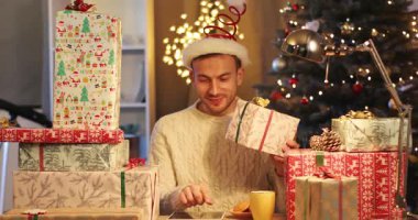 Komik Noel Baba şapkalı, gülümseyen yakışıklı adam güzel renkli hediye kutularını sayıyor. Genç adam Noel ağacının yanında dijital tablet kullanıyor. Kapalı.