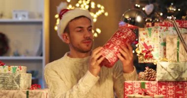 Komik Noel Baba şapkalı çekici genç adam renkli ambalaj kağıdıyla hediye kutularını sayıyor. Adam Noel ağacının yanında dijital tablet kullanıyor. Kapalı.