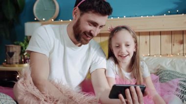 Oldukça neşeli bir baba ve kızının videolar izleyip eğlenirken çekilmiş bir portresi. Yakın plan küçük kız ve babası evde pembe giyinmiş sihirli periler oynuyorlar..