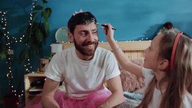 Küçük kız, mutlu baba yüzüne fırçayla göz farı sürüyor. Neşeli gülümseyen baba ve kız birlikte oynuyorlar. Ev partisinde sihirli peri gibi giyiniyorlar..