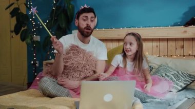 Neşeli güzel baba ve kızı dizüstü bilgisayardan çizgi film izlerken sihirli numaralar yapıyor. Evde yatakta oturan periler gibi giyinmiş genç bir adamla küçük kızının aile portresi..