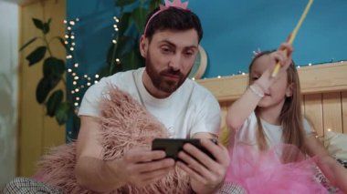 Mutlu baba-kız sihirli perileri giydirip birlikte oynayarak akıllı telefondan video izliyorlar. Sihirli değnekle babasıyla yatakta el sallayan tatlı kız. doğum günü partisi.