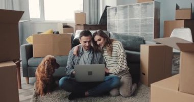 Genç bir çift, taşınan kutuların arasında oturmuş, dizüstü bilgisayarları üzerinde çalışıyor ve köpekleri yakınlarda geziniyor.