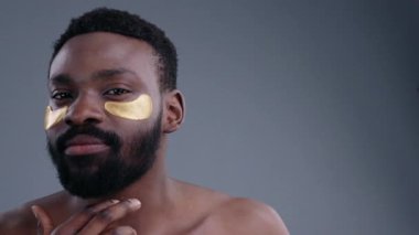 Göz altı altın bantları olan çekici Afrikalı genç adam gri arka plandaki kameraya bakıyor..