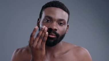 Yakışıklı, üstsüz, Afro-Amerikan bir adam banyoda yüz bakımı için sivrisinek kremi kullanıyor. Siyah Maler modelinin portresi gri arkaplanda aynaya bakıyor..