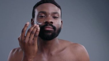 Gri arkaplandaki kameraya bakarak yüz kremi süren yakışıklı, duyarlı siyahi bir adamın yakın çekimi. Erkek güzellik ve bakım konsepti.