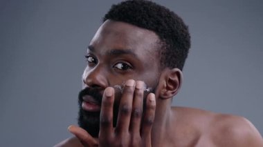 Çekici bir Afrikalı gencin portresi. Nemlendirici bir yüz kremi kullanıyor. Yüzüne nazikçe dokunuyor. Yakından bakınca yakışıklı bir adamın kameraya gülümseyen bir krema kavanozunu göstermesi..