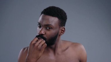Beyaz dişlerini kamerada fırçalayan ve gösteren yakışıklı Afro-Amerikalı adamın yakın çekimi. Gri arka planda parlak bir gülümsemesi olan neşeli, siyah gömleksiz bir adamın portresi..